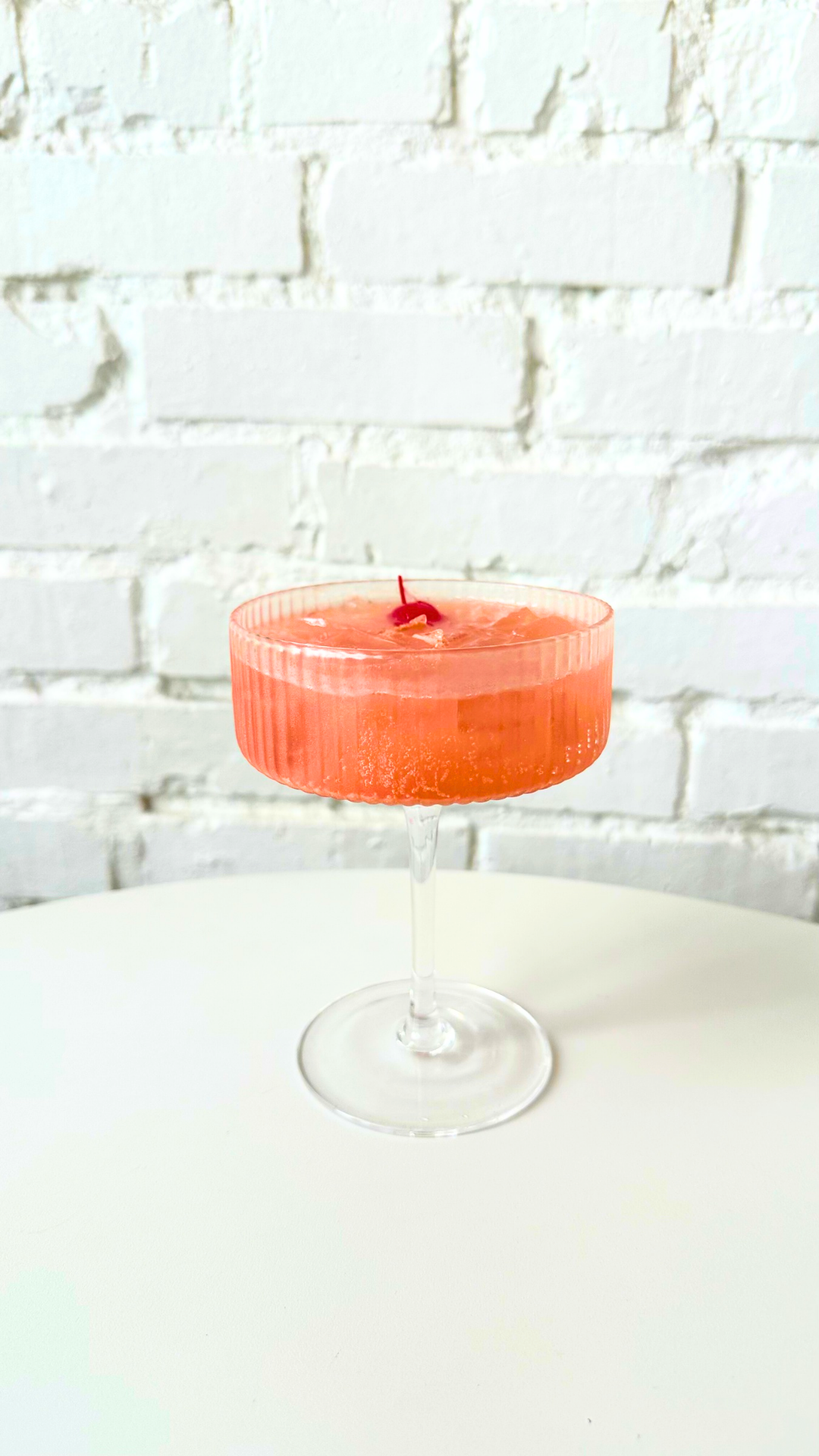A pink Dirty Shirley Kava Nectar mocktail over ice in a ribbed coupe glass, topped with a maraschino cherry.