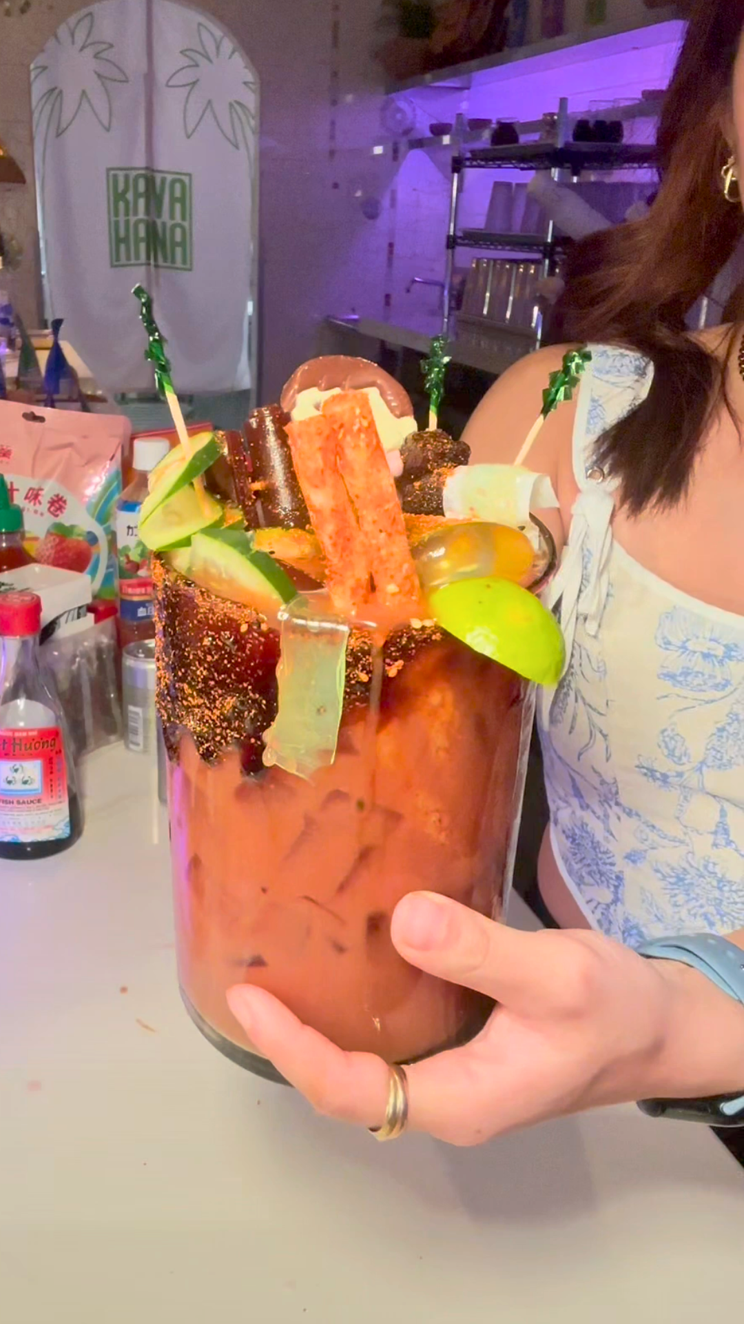Holding a tall Fish Sauce Kava Nectar Michelada over ice with a chili-spiced rim, garnished with cucumber, lime and candy on skewers, at the Kavahana bar.