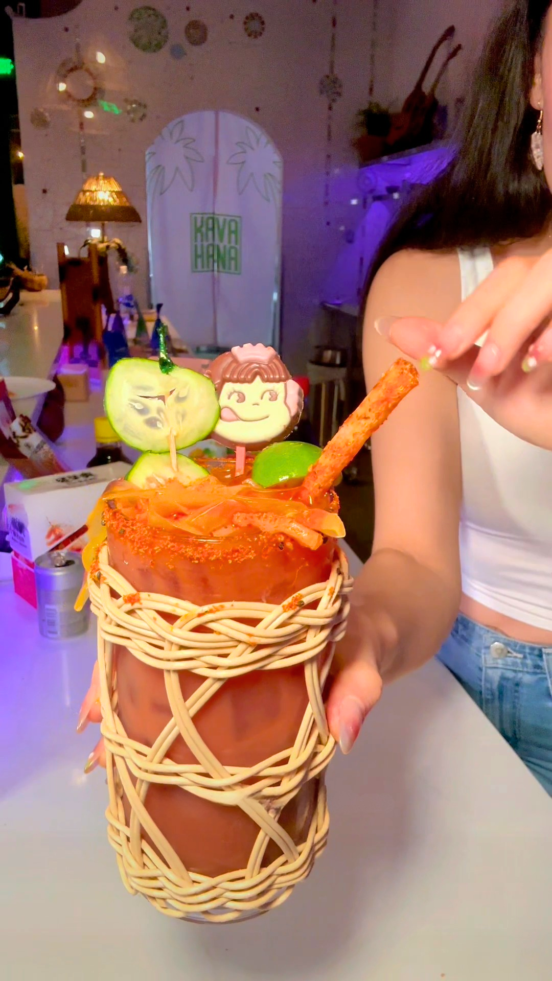 A Japanese Kava Nectar Michelada over ice with a seven spice rim, cucumber and lime garnish, in a woven glass holder.
