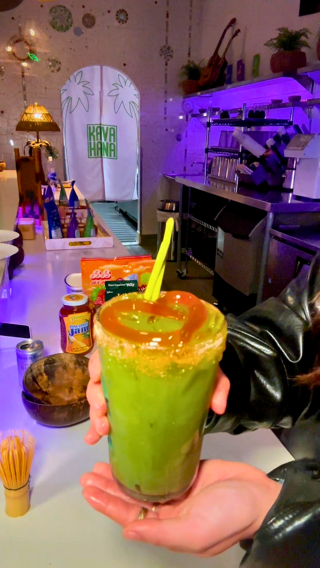 Holding a tall green Matcha Kava Nectar Michelada with a sugared rim drizzled with caramel and a yellow straw, at the Kavahana bar. 