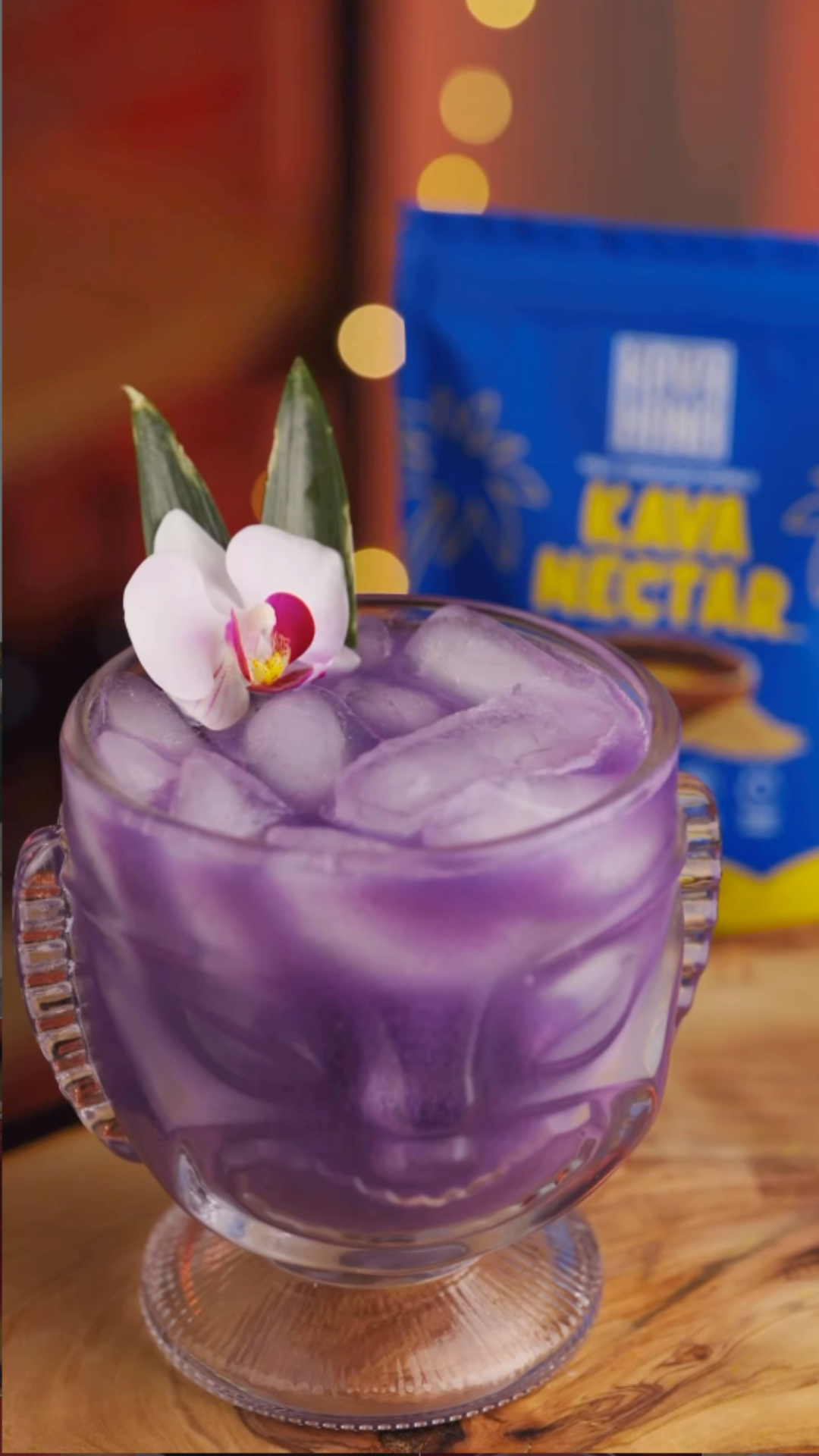 A purple ube piña Kava Nectar mocktail in a tiki glass over ice, garnished with an orchid and pineapple fronds, beside a Kava Nectar Relax Edition bag.